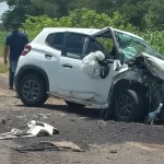 Veículo branco com a frente destruída após o grave acidente na BR-376 em Guairaçá, parado às margens da rodovia ao lado de um milharal, com pessoas ao fundo.