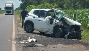 Veículo branco com a frente destruída após o grave acidente na BR-376 em Guairaçá, parado às margens da rodovia ao lado de um milharal, com pessoas ao fundo.