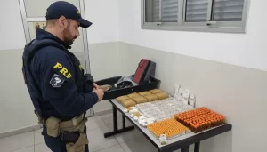 Policial rodoviário federal em perfil observando a apreensão de medicamentos pela PRF organizada sobre uma mesa, com centenas de frascos de emagrecedores e cartelas de comprimidos