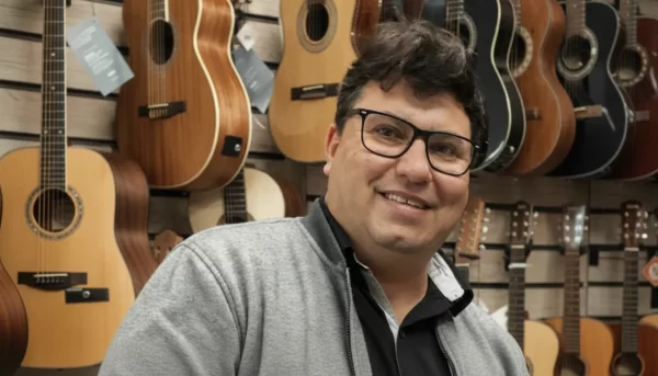 Retrato restaurado do empresário de Umuarama, Ricardo Ferri da Silva, sorrindo e usando óculos em frente a uma parede com diversos violões em sua loja.