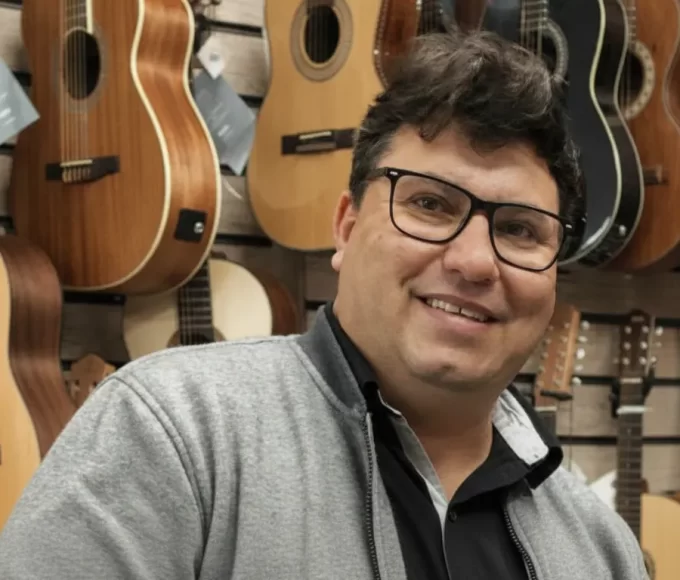 Retrato restaurado do empresário de Umuarama, Ricardo Ferri da Silva, sorrindo e usando óculos em frente a uma parede com diversos violões em sua loja.