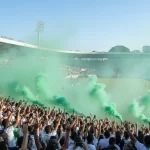 Grande multidão de torcedores com seus Maringá FC ingressos celebrando nas arquibancadas do Estádio Willie Davids, em meio a uma densa e vibrante nuvem de fumaça verde.