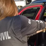 Agente da PCPR Umuarama em ação, vista de costas utilizando colete tático da Polícia Civil e apontando arma para dentro de um veículo durante operação