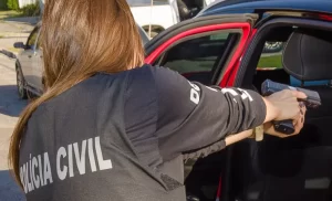 Agente da PCPR Umuarama em ação, vista de costas utilizando colete tático da Polícia Civil e apontando arma para dentro de um veículo durante operação