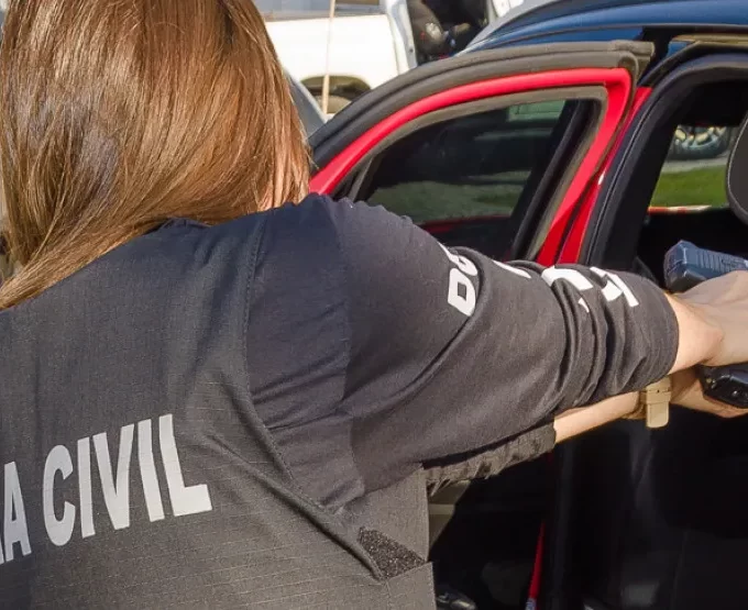 Agente da PCPR Umuarama em ação, vista de costas utilizando colete tático da Polícia Civil e apontando arma para dentro de um veículo durante operação