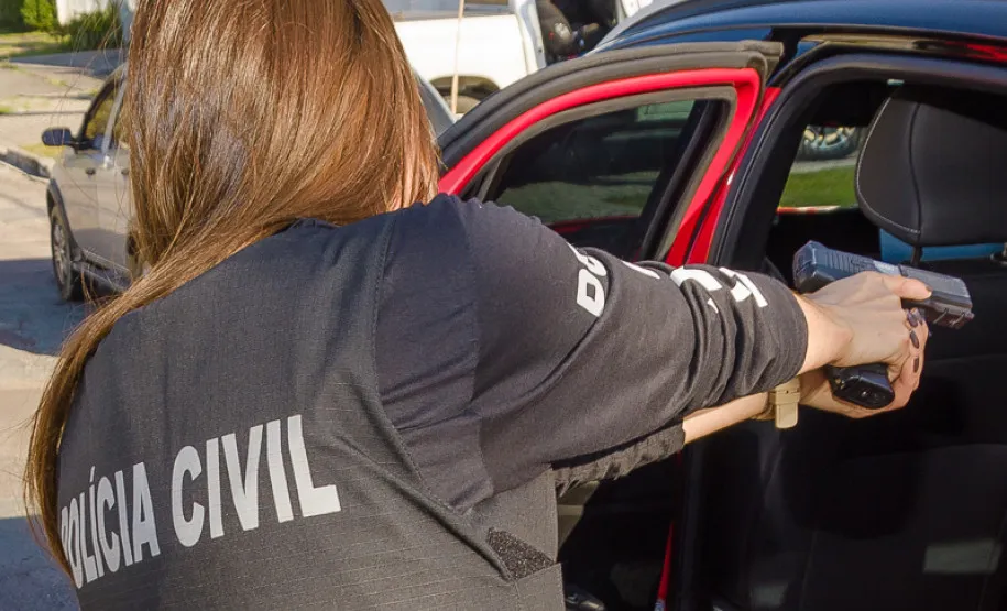 Agente da PCPR Umuarama em ação, vista de costas utilizando colete tático da Polícia Civil e apontando arma para dentro de um veículo durante operação